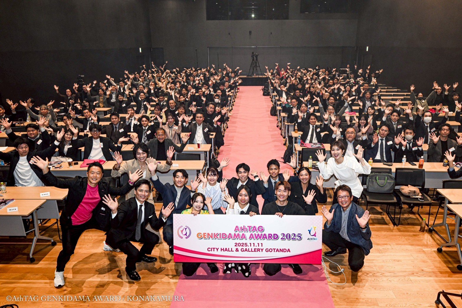 2025年大会の集合写真。CITY HALL & GALLERY GOTANDA の客席いっぱいの参加者が手を挙げ、前列のファイナリストと審査員が大会名の入った横断幕を掲げている。